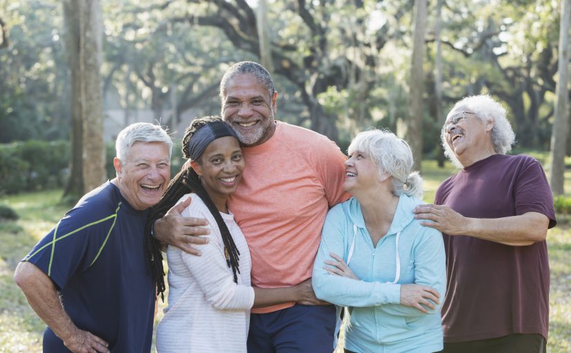 A group of five multi-ethnic seniors standing together in a park wearing casual clothing. The African-American couple in the middle are smiling and the others are laughing.