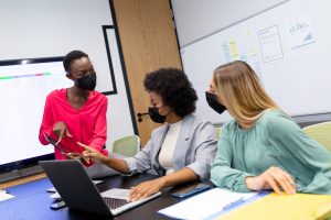 Three women, one with a mobile device, one with a laptop computer, and one with a folder of papers, discuss information displayed on the mobile device