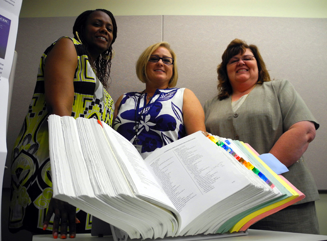 Medical coders Celia Dickens, Tyringa Crawford, and Holly Lambert pose behind a manual. They are part of a select group; around the world there are only a few mortality coders.