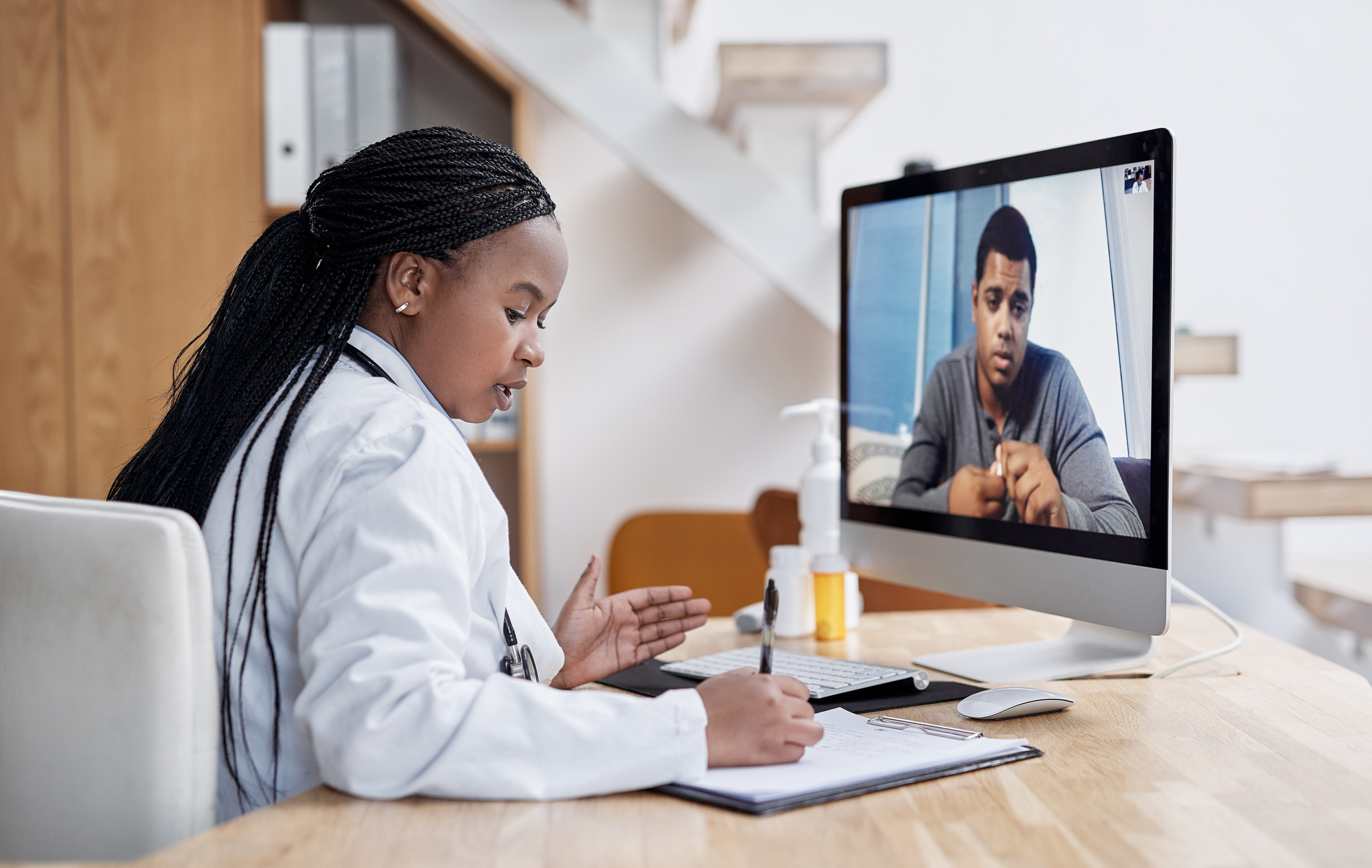 Shot of a young doctor writing notes during a video call with a patient on a computer.