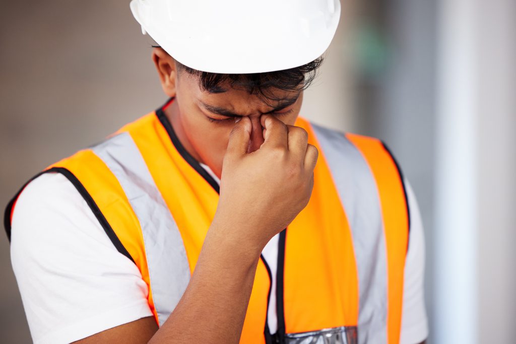 A young construction worker holding bridge of nose with eyes closed. 