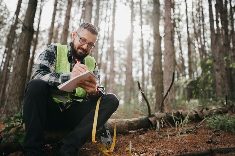 A male worker in a yellow safety vest is writing on a note pad alone in a forest