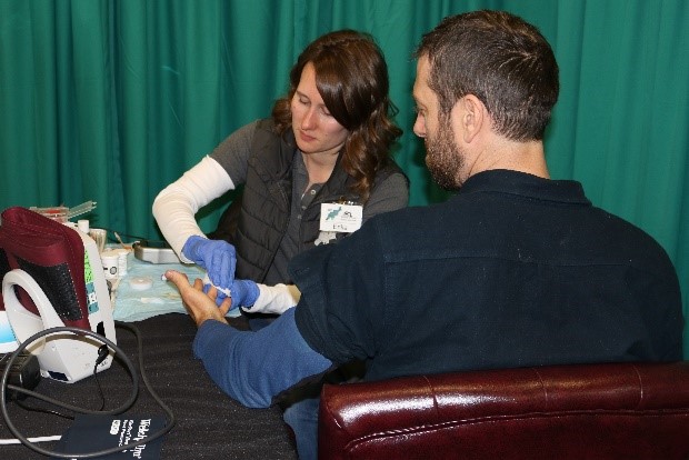 A female doctor providing a cholesterol and glucose screening to a male logger.