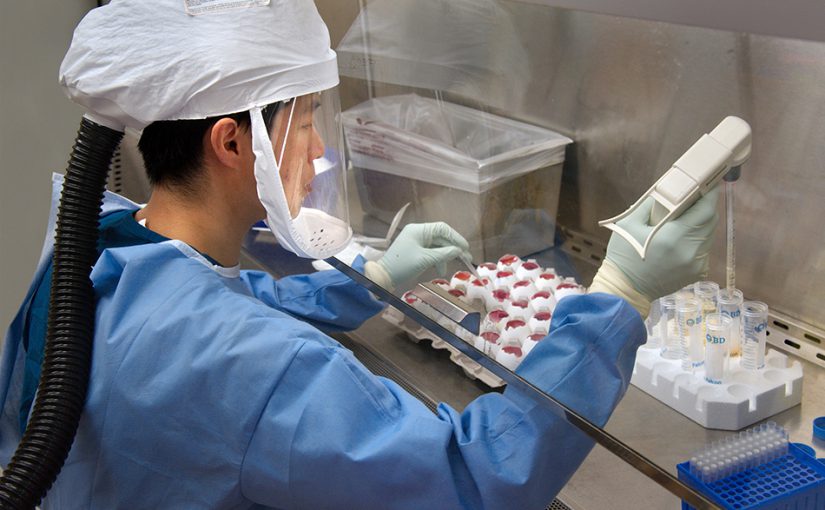 A CDC scientist is wearing a protective airtight suit, equipped with a helmet and face mask. She is seated, pipetting specimens in a laboratory.