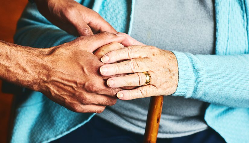 A male caregiver holds the hand of an older woman.