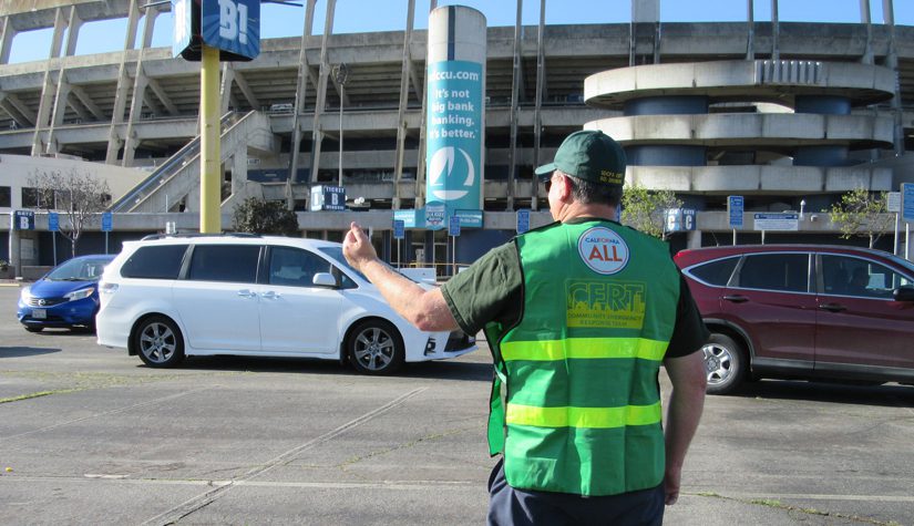 A man in a green vest and hat directs traffic in parking lot.
