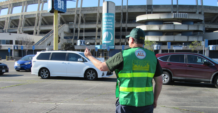 A man in a green vest and hat directs traffic in parking lot.