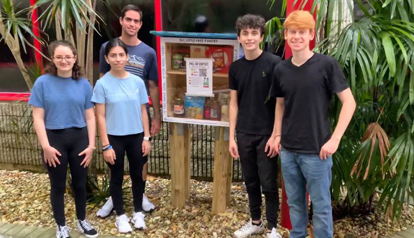 Five teenagers stand beside a freestanding wooden container filled with food. A label on the box says "TLC little free pantry"