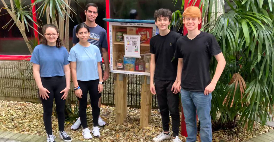 Five teenagers stand beside a freestanding wooden container filled with food. A label on the box says "TLC little free pantry"