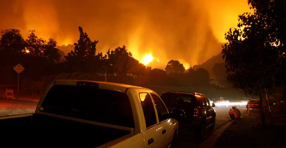 A line of vehicles parked along the side of a road at night. A wildfire burns in the distance.