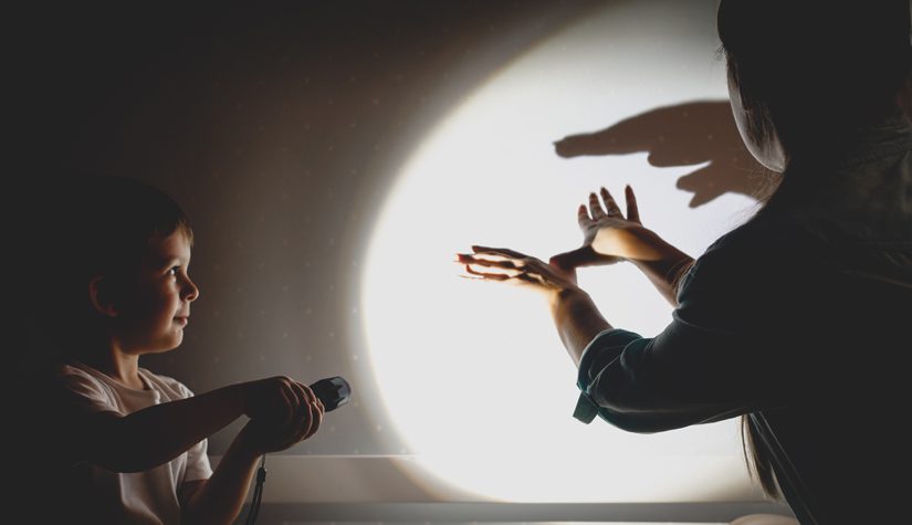 A young woman and boy make hand shadow puppets using a flashlight against a white wall.