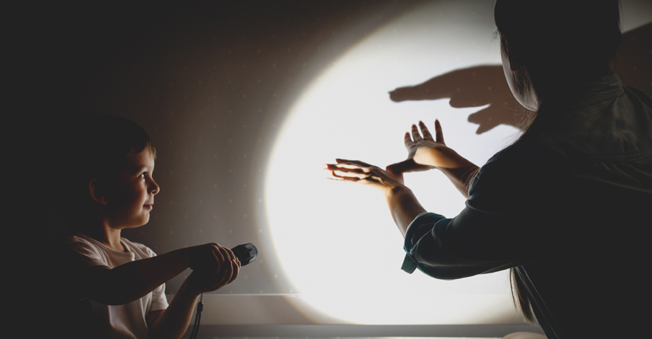 A young woman and boy make hand shadow puppets using a flashlight against a white wall.