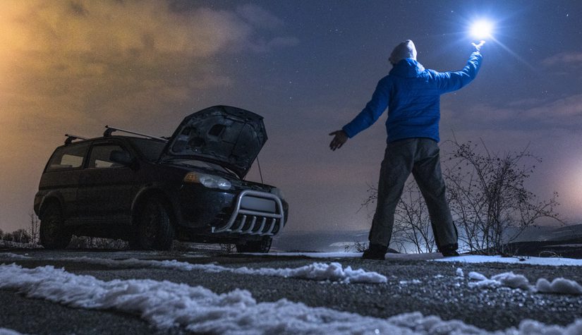 A person holding up a flashlight. Their car is broken down on the side of a snowy road.