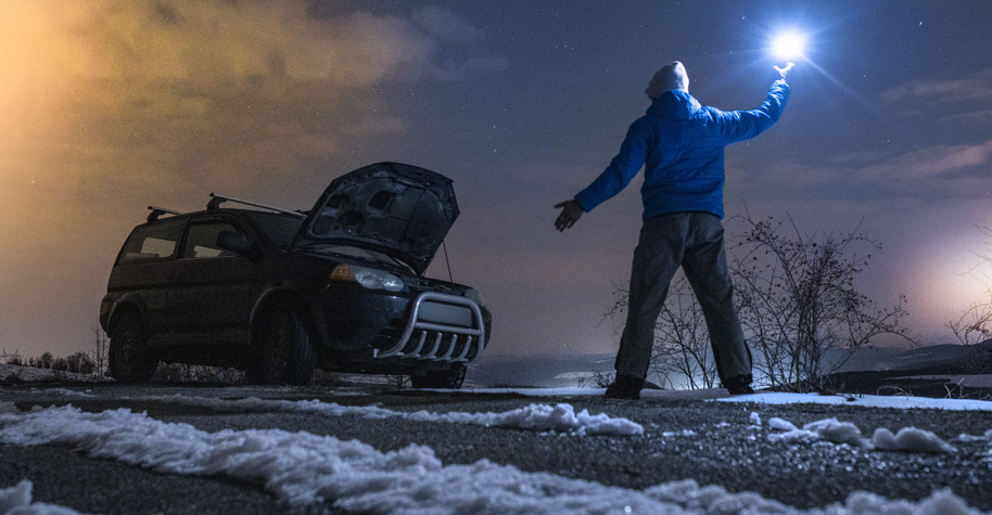 A person holding up a flashlight. Their car is broken down on the side of a snowy road.