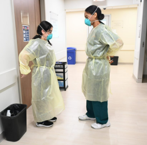 Two female healthcare workers in hallway prepare to remove their personal protective equipment.