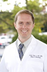 Dr. Cosby Stone in white medical coat, shirt, and tie. Trees faded in background.