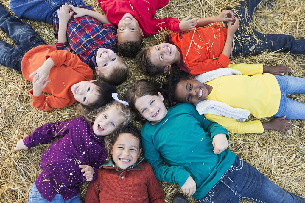 A group of eight multi-ethnic children, 4 to 6 years old, lying on their backs on hay, in a circle with heads in the middle, smiling at the camera.