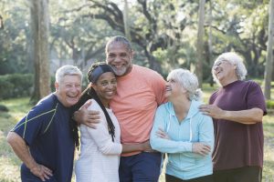 A group of five multi-ethnic seniors standing together in a park wearing casual clothing. The African-American couple in the middle are smiling and the others are laughing.