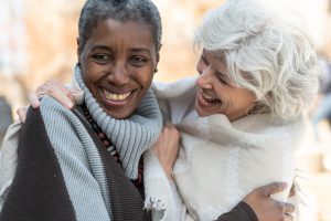 Two senior friends laugh affectionately together. The women are standing outside on a sunny but cool day. They are dressed in casual sweaters. One woman is of African descent and the other is Caucasian.