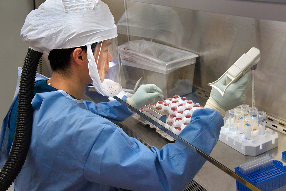 A CDC scientist is wearing a protective airtight suit, equipped with a helmet and face mask. She is seated, pipetting specimens in a laboratory. 