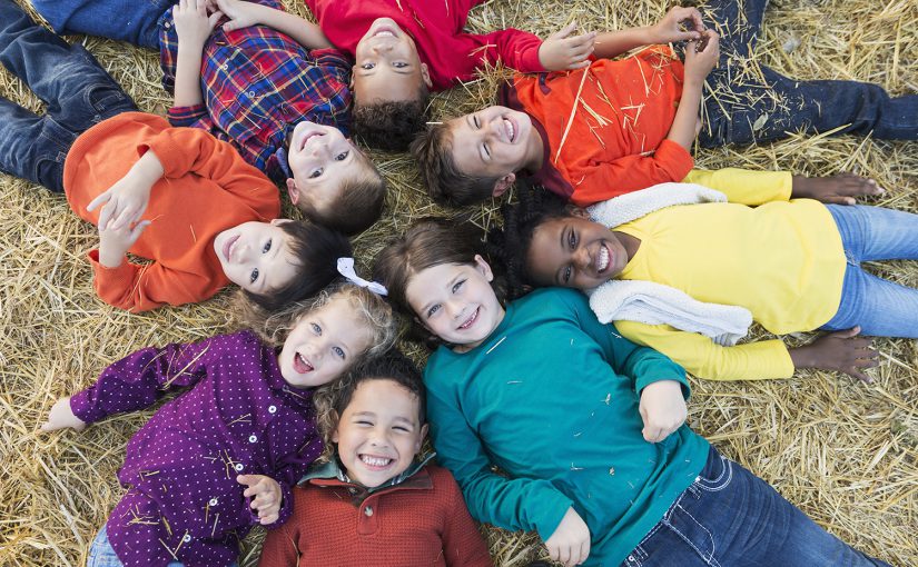 A group of eight multi-ethnic children, 4 to 6 years old, lying on their backs on hay, in a circle with heads in the middle, smiling at the camera.