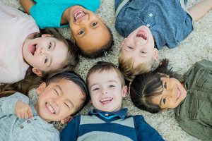 Diverse group of young children laying down in a circle with their heads togethers all smiling at the camera.