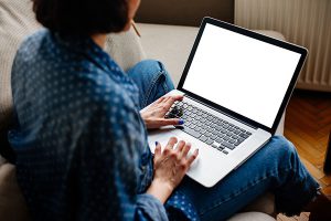 A woman with a blue and white shirt and blue jeans sitting on a couch looking at a blank laptop screen.